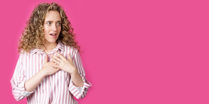 The woman holds her hands on her heart, wearing a striped shirt, expresses gratitude, has a friendly expression on her face. Isolated on pink background
