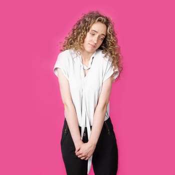 Young woman with curly hair, smiling broadly, squeezes her shoulders. Isolated on pink background