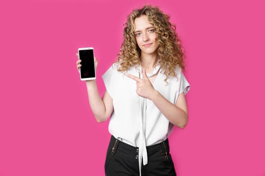 Look at this cell phone. Contented happy woman, pointing her index finger at a blank screen, shows a modern device. Isolated on a pink background. Technology concept