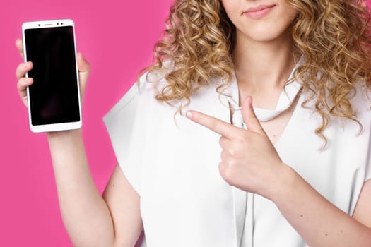 Look at this cell phone. Contented happy woman, pointing her index finger at a blank screen, shows a modern device. Isolated on a pink background. Technology concept