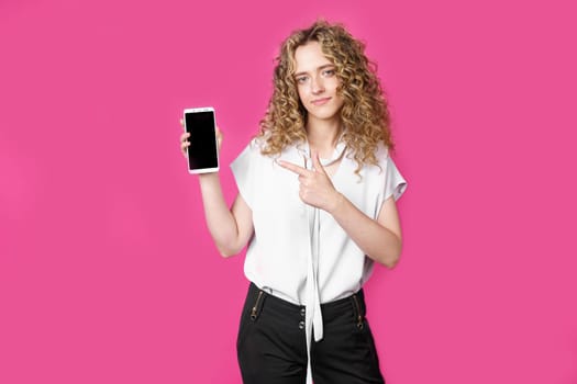 Look at this cell phone. Contented happy woman, pointing her index finger at a blank screen, shows a modern device. Isolated on a pink background. Technology concept