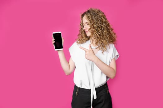 Look at this cell phone. Contented happy woman, pointing her index finger at a blank screen, shows a modern device. Isolated on a pink background. Technology concept