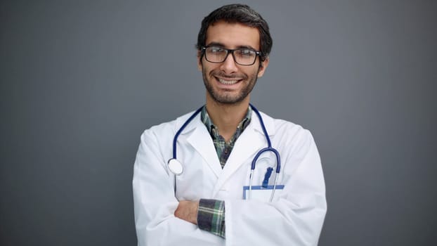 Portrait of positive male doctor posing with crossed arms and smiling at camera over grey studio background