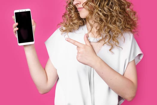 Look at this cell phone. Contented happy woman, pointing her index finger at a blank screen, shows a modern device. Isolated on a pink background. Technology concept