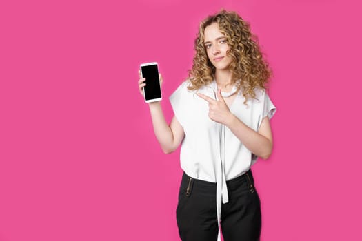 Look at this cell phone. Contented happy woman, pointing her index finger at a blank screen, shows a modern device. Isolated on a pink background. Technology concept