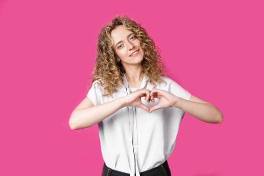 Portrait of a beautiful woman who shows a heart gesture on her chest, passionately, expresses love for a loved one. Isolated pink background.