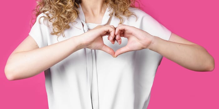 Portrait of a beautiful woman who shows a heart gesture on her chest, passionately, expresses love for a loved one. Isolated pink background.