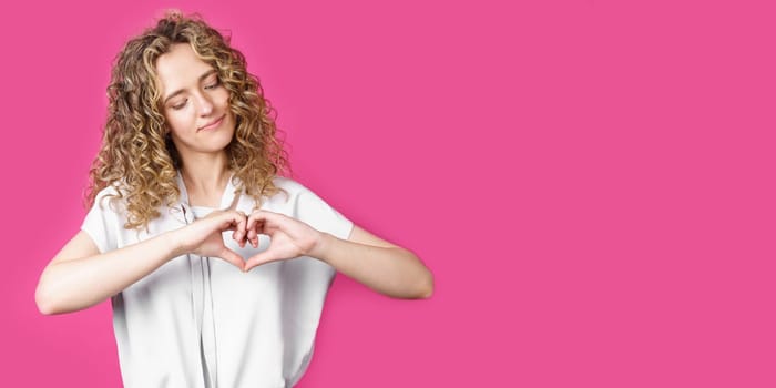 Portrait of a beautiful woman who shows a heart gesture on her chest, passionately, expresses love for a loved one. Isolated pink background.
