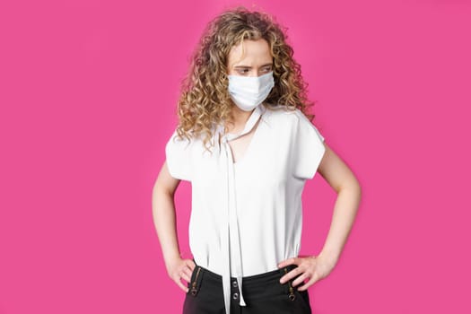 A young woman with a medical mask on her face is standing with her hands on her waist. Isolated pink background