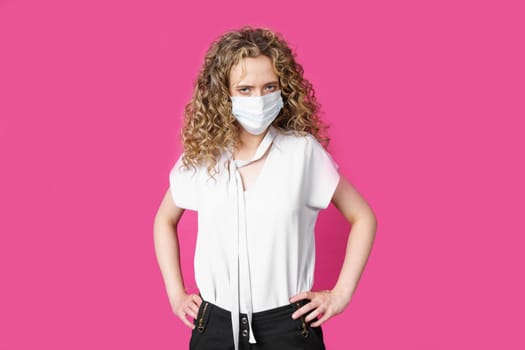 A young woman with a medical mask on her face is standing with her hands on her waist. Isolated pink background