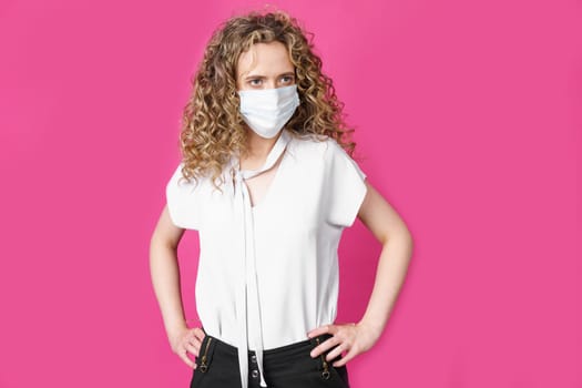 A young woman with a medical mask on her face is standing with her hands on her waist. Isolated pink background