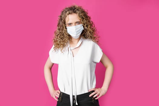 A young woman with a medical mask on her face is standing with her hands on her waist. Isolated pink background