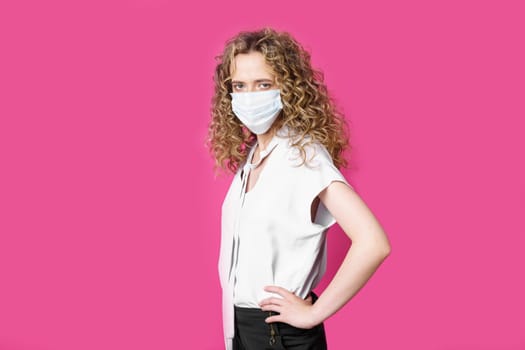 A young woman with a medical mask on her face is standing with her hands on her waist. Isolated pink background