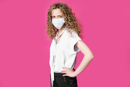 A young woman with a medical mask on her face is standing with her hands on her waist. Isolated pink background