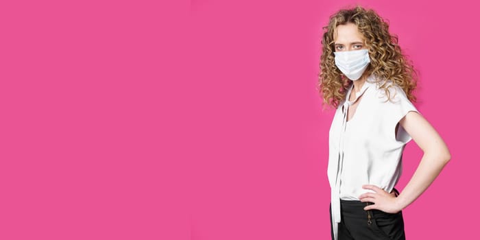 A young woman with a medical mask on her face is standing with her hands on her waist. Isolated pink background