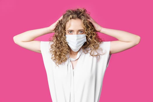 A young woman with a medical mask on her face holds her head with her hands. Isolated pink background