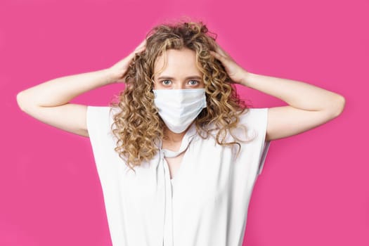 A young woman with a medical mask on her face holds her head with her hands. Isolated pink background