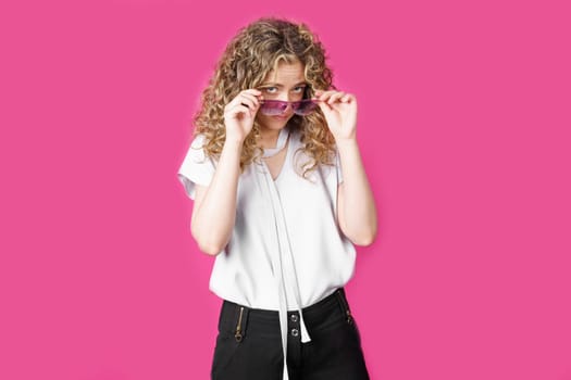 A young woman holds glasses with her hands and looks from under them. Isolated pink background