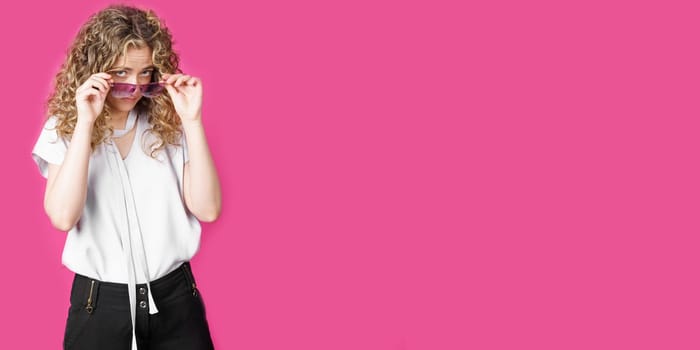 A young woman holds glasses with her hands and looks from under them. Isolated pink background