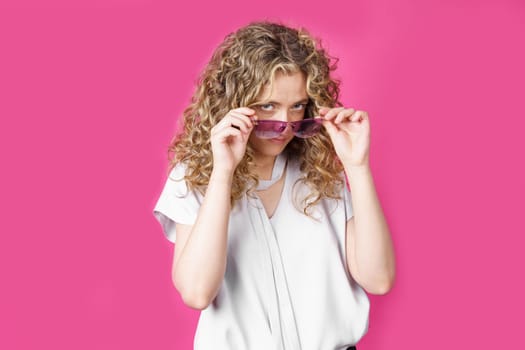 A young woman holds glasses with her hands and looks from under them. Isolated pink background