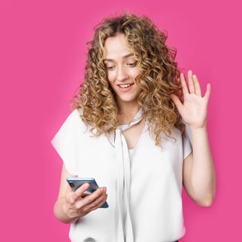 Young woman communicates through the phone by video link, expressing emotions. Female portrait. Isolated on pink background