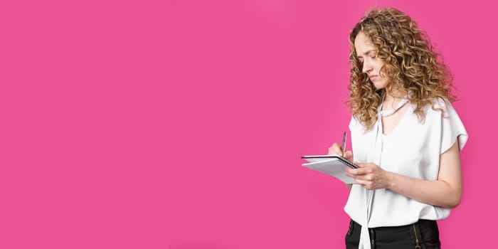 A charming woman makes entries in the diary. Isolated on a pink background.