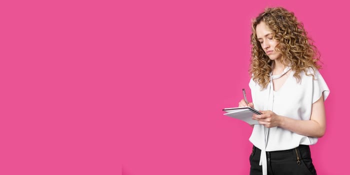 A charming woman makes entries in the diary. Isolated on a pink background.