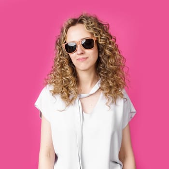 Young woman with curly hair, smiling broadly, squeezes her shoulders. Isolated on pink background