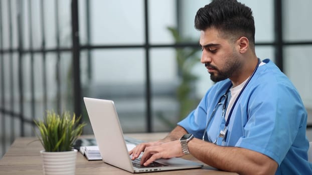 Young male doctor works on laptop computer at table in medical office.