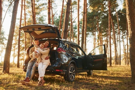 Leaning on the car. Happy family of father, mother and little daughter is in the forest.