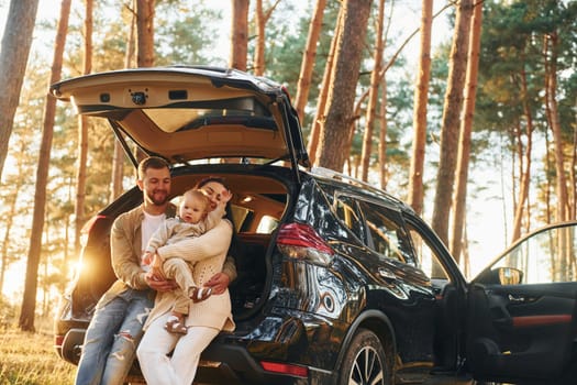Leaning on the car. Happy family of father, mother and little daughter is in the forest.