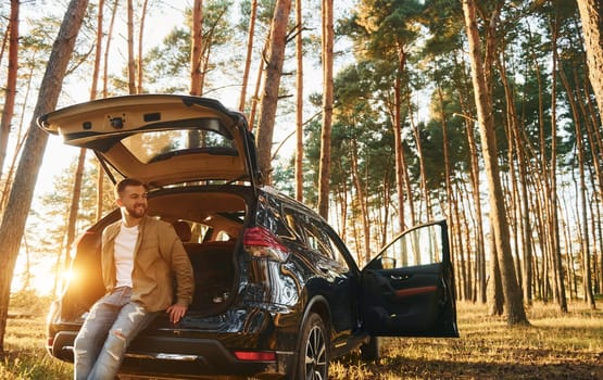 With his car. Man in jeans is outdoors in the forest with his black colored automobile.