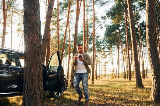 Near the car. Man in jeans is outdoors in the forest with his black colored automobile.