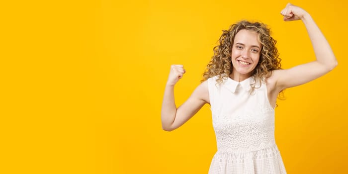 A strong woman with curly hair, a toothy smile, raises her arms and shows her biceps. Models on a yellow background. Look at my muscles