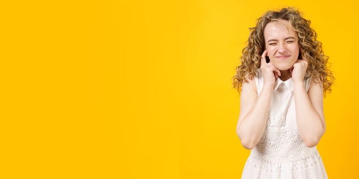 Portrait of a young woman who covered her ears with her fingers. Isolated on yellow background