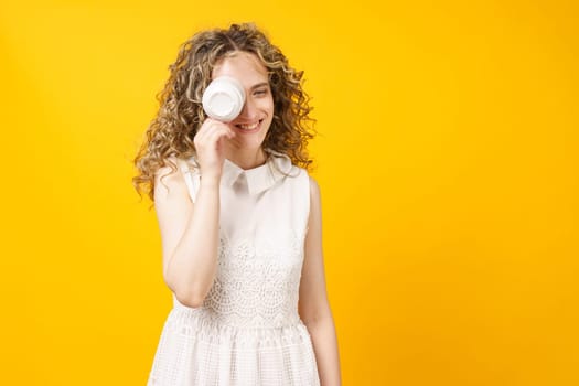 Young woman is covering her face with a cup and smiling. Female portrait. Isolated on yellow background