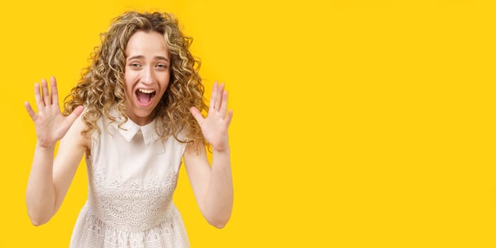 A curly-haired woman raises her palms in joy, glad to receive an amazing gift from someone, screams loudly, wearing a striped shirt. Isolated on a yellow background.