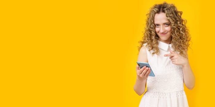 Portrait of a young smiling woman who points to her cell phone. Female portrait. Isolated on yellow background