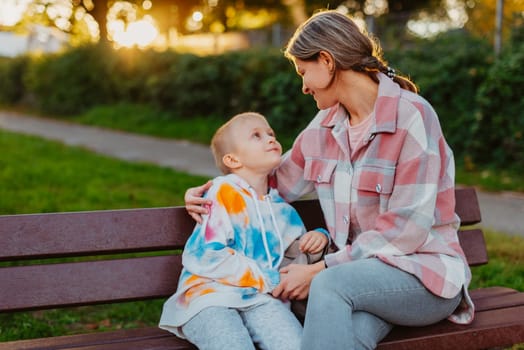 mother and son sit on a park bench in the rays of the setting sun. the concept of a family. Mother's Day. beautiful girl (mother) with a boy (son) in the park in the park are sitting on a bench at sunset.