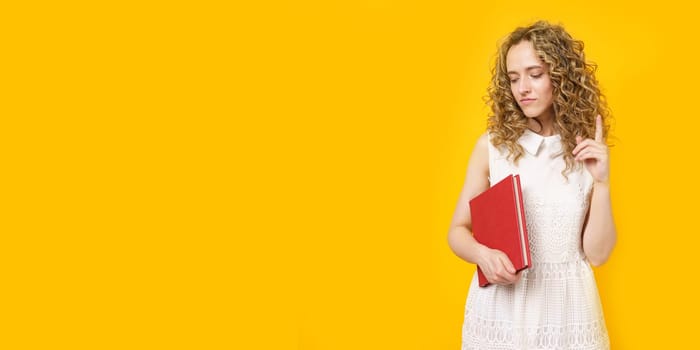 Portrait of a young smiling woman who holds a book in her hands and points her finger to the side. Female portrait. Isolated on yellow background