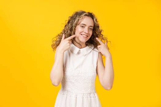 Portrait of a young smiling woman showing her smile with her fingers. Female portrait. Isolated on yellow background