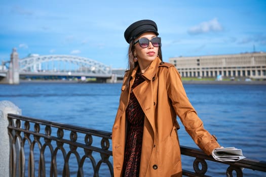 A female portrait of European appearance on the embankment on a sunny spring day with a newspaper in her hand. Beautiful brunette woman in sunglasses and in a brown jacket and black cap