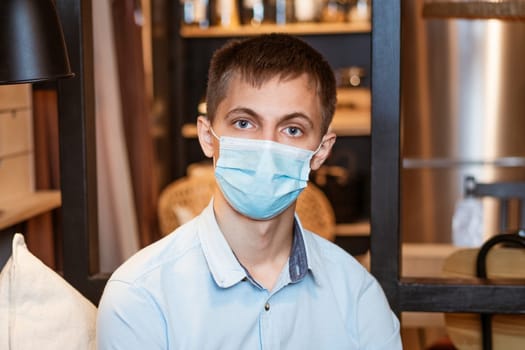 a young guy of Caucasian ethnicity in a shirt and a protective mask sits on the couch alone at home. Concept of being at home during global quarantine