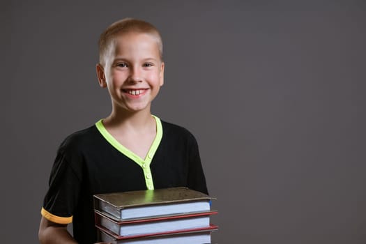 Cheerful caucasian boy in a black t-shirt holds a stack of books on a gray background. Education concept