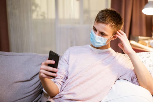 young man of European appearance in casual clothes, at home on the couch in a medical mask communicates by video communication on the phone, the concept of remote online communication on the Internet