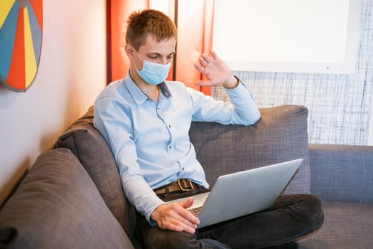 a young guy of European appearance in a light shirt, sits in a protective medical mask and works on the couch via video communication, remote work during a global pandemic