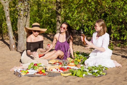 Cheerful girlfriends are having fun in summer at picnic. cheerful company beautiful girls. Young caucasian women wearing sunglasses and drinking wine drinking wine and eating fresh fruits in nature