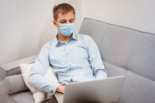 a young man of Caucasian appearance in a blue shirt and a protective mask with a laptop on the couch works at home. Remote work in quarantine