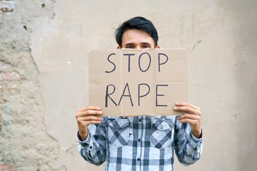 Young man holding cardboard with the inscription stop rape. Guy of Caucasian appearance against violence holds a poster with a protest at the demonstration against the background of the wall