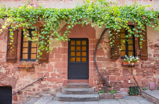 front of a house built of red bricks with a large green grape plant above the door and shuttered windows